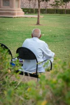 Old man isolated sitting alone at iron chair at park from back at evening 库存照片