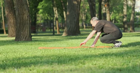 Old man lies down on a mat spread grass city park. Beginning of yoga, physical Stock Footage 243908210