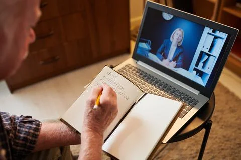 Old man making notes in notebook while sitting in front of laptop Foto stock