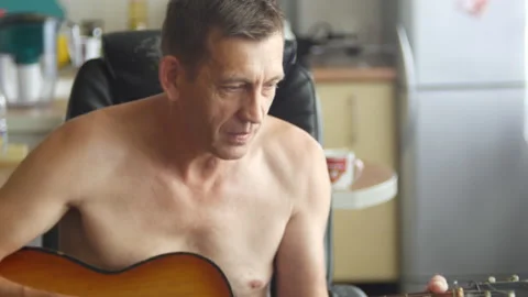 Old man is playing on a six string acoustic guitar sitting on chair in the Stock Footage 136729458