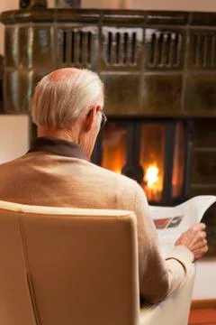Old man reading newspaper while sitting by a fireplace in a living room Фото
