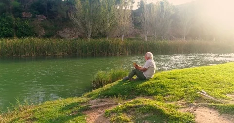Old man reading sitting on the grass by the river Stock Footage 124381993
