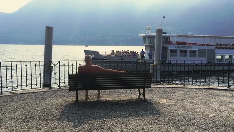 Old man rests on a bench on the como lake while ferry is passing by Stock Footage 118649876