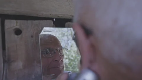 Old man shaves beard looking at mirror reflection on fence Stock Footage 249957490
