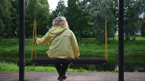 Old man sits alone on park swing. Swings gently above grassy pond. Trees fr.. Stock Footage 340039919
