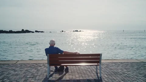Old man sitting on a bench overlooking sea or ocean enjoying sunny day outside Stock Footage 304460524