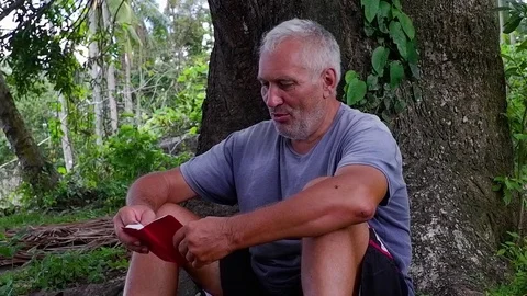 An old man sitting under the tree while reading a book. Video stock 122476001