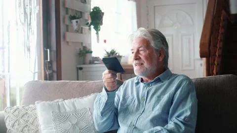 Old man smiling sitting on the sofa in the living room holding phone, enjoying u Stock Footage 151326356
