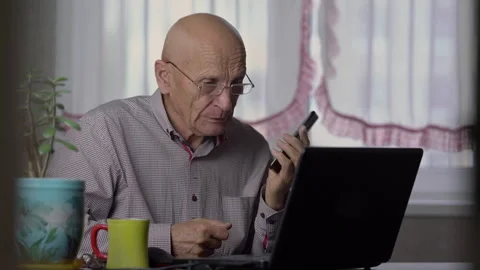 Old man talks to master on phone and presses keys on laptop Stock Footage 242197741