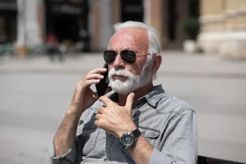 Old man use internet application to speak on smartphone, seating on bench,  s Stock Photos