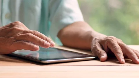 Old man using tablet with right index finger on light brown wooden table surface Stock Footage 80903699