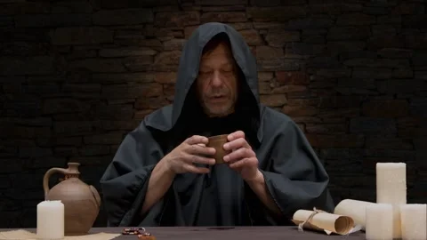 An old monk in a black robe pours wine into a goblet from a jug and drinks while Stock Footage 274455956