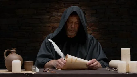 An old monk in a black robe writes with a quill in a scroll of parchment, sittin Stock Footage 274455872