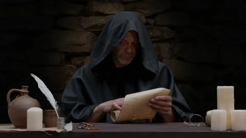 An old monk opens a scroll and reads, sitting at a table against a stone wall,  Video stock 274455835