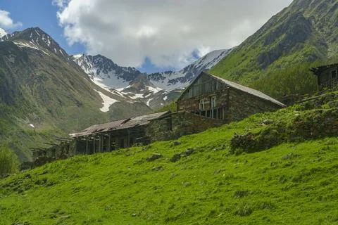 Old mountain farmstead on green slope beneath snowy Caucasus ridges Stock Photos
