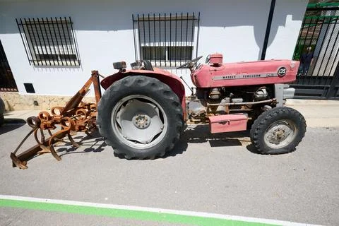 Old red tractor with plow attachment parked on a sunny village street Stock Photos