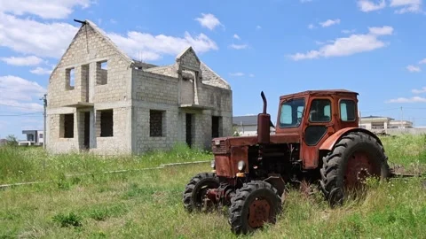 Old red tractor standing in overgrown grass beside unfinished brick house in Stock Footage 337937529