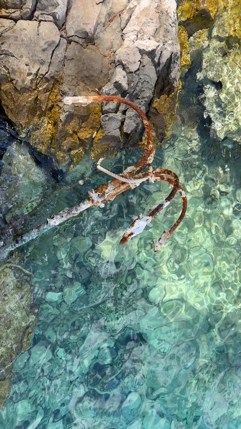 Old rusty anchor resting on rocky shore in crystal clear turquoise sea water Stock Footage 314945168