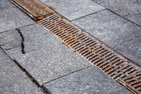 Old rusty drainage grate on granite pavement with ruined cracked tiles. Stock Photos