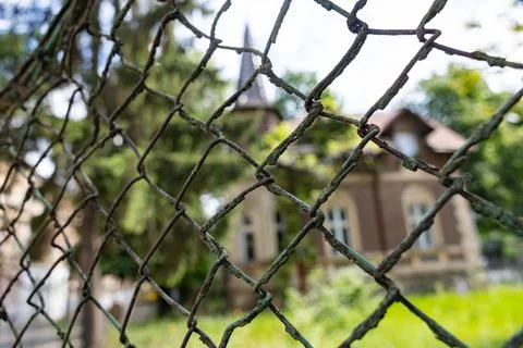 Old rusty metal wire mesh fence close-up with a blurred historic house and .. Stock Photos