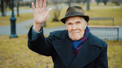 Old senior man waves his hand, greets, says Hello. He is happy sitting in park. Stock-Footage 129574946