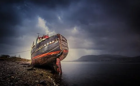 Old shipwreck rests on the shore under a cloudy sky at dusk near a tranquil body Stock-Fotos
