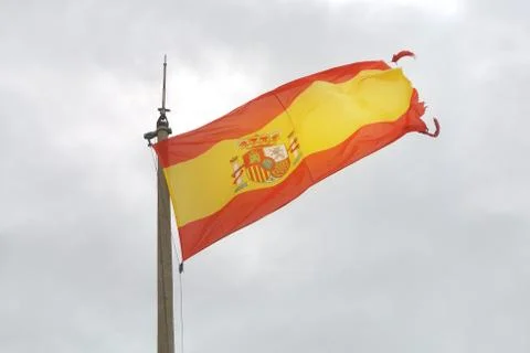 Old Spanish flag on a pole, undulating in the wind with clouds at the backgro Stock Photos
