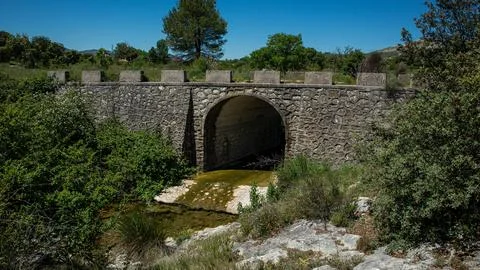 Old stone arch bridge spans small river in Vall de Alcala under clear blue .. Stock Photos