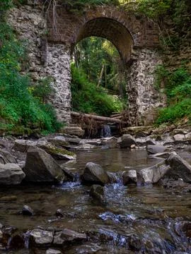 Old stone arched destroyed bridge over small river in a mountain forest Stock Photos