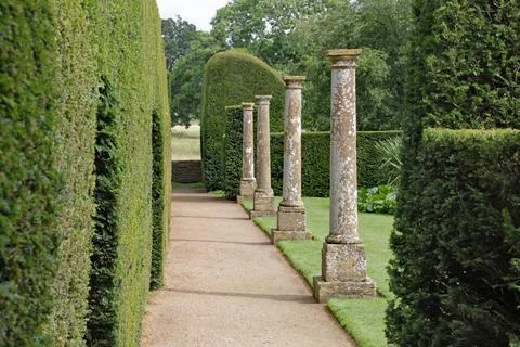 Old stone columns stand by a hedge with arch shaped doorways cut in it at an Stock Photos