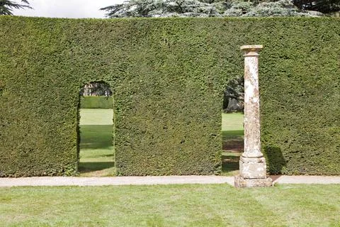 Old stone columns stand by a hedge with arch shaped doorways cut in it at an Stock Photos