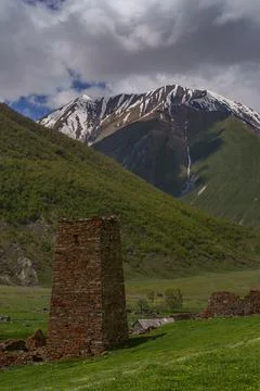 Old stone tower beneath steep green slope and snowy summit Foto stock
