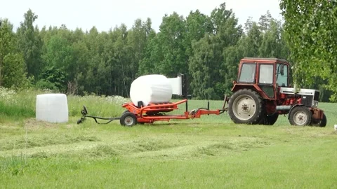 Old tractor pulls a bale wrapper across a cut grass field Video stock 337257020