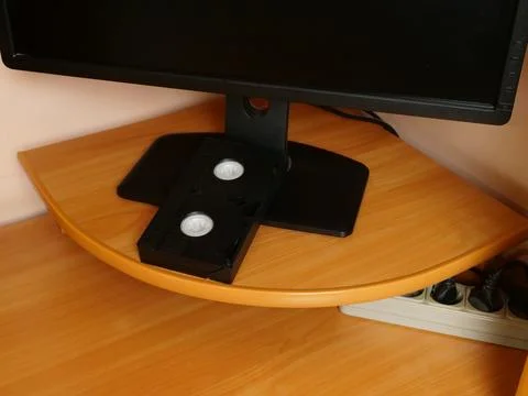 Old video cassette placed under computer monitor on wooden corner desk in hom Stock Photos