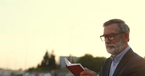 Old wise Caucasian gray-haired man in glasses walking and reading book outdoors Video stock 139931588