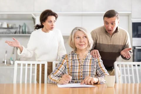 Old woman compiling heritage document while middle-aged man and woman competing Stock Photos