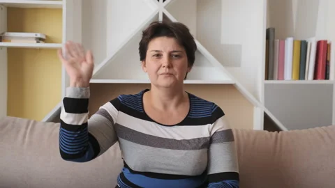 Old woman looking at camera sit on sofa alone at home and waving hello. Stock Footage 134870817