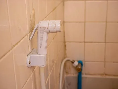Old worn white plastic bidet hand sprayer mounted on beige ceramic bathroom.. Stock Photos