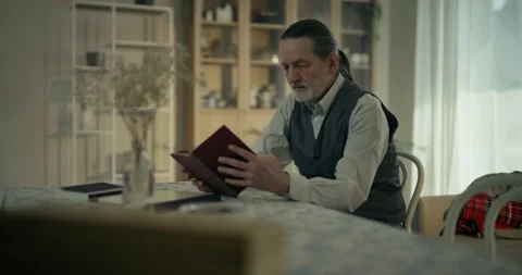 Older bearded man writing on papers at a table in a cozy home interior Stock Footage 336286397
