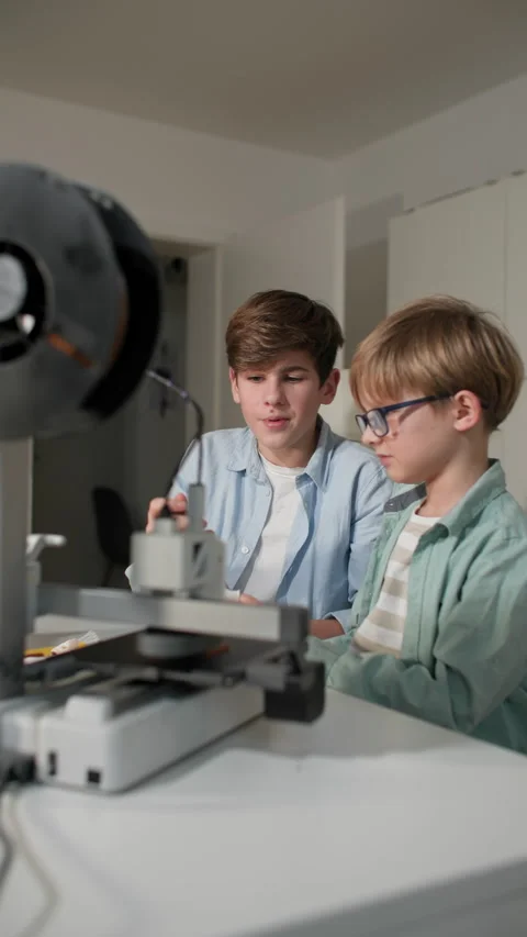 Older boy explains to a younger boy wearing glasses how a 3D printer works Stock Footage 335390560
