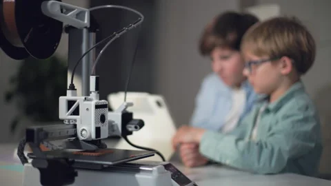Older of the children starts up the 3D printer in the foreground of shot Stock Footage 335376792