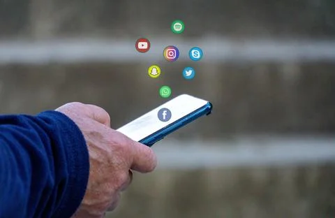 Older female hand holding a blue smartphone with floating social media icons Stock Photos