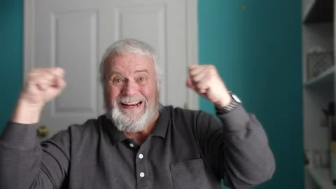 An older man expresses joy and excitement while sitting in a room. He raises Stock Footage 338058844