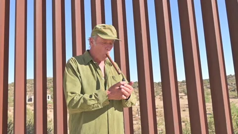 Older man in front of US Mexico border fence lights a cigar. Stock Footage 168721393