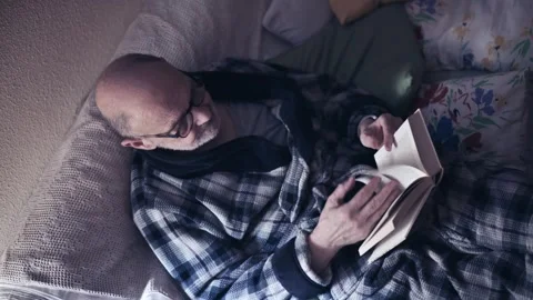 Older Man Reading a Book on a Couch Wrapped in a Plaid Blanket at Home Stock Footage 318122697