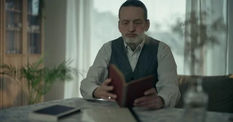 Older man reading a book at home beside a bright window in soft daylight Видео 336279892