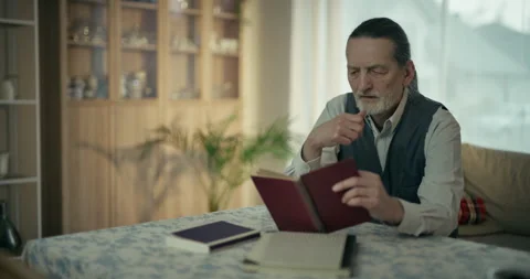 Older man reading a book at a table in a cozy sunlit living room Stock Footage 336276292