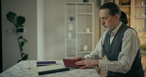Older man reading a red document at a dining table in a home interior Stock Footage 336287751