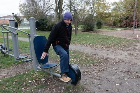Older man sits on exercise machine in local park surrounded by trees Stock Photos