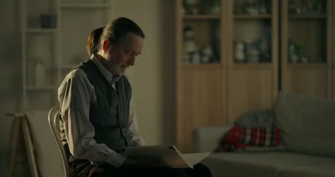 Older man sitting quietly in a dim living room with soft window light Stock Footage 336276544